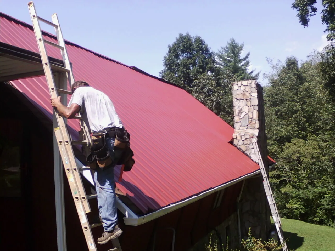 Roofer on a ladder beside a red metal roof and stone chimney — Soffit & Fascia Repair by Roofing Brothers in Durant